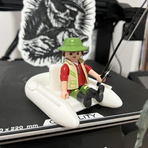 May include: A plastic figurine of a fisher wearing a green hat, tan vest, and red shirt, sitting in a white inflatable boat while holding a black fishing rod. The figure is positioned on a black surface with the text CREALITY visible.