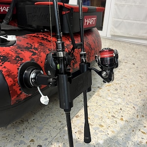 May include: A black plastic fishing rod holder mounted on a red and black camouflage inflatable boat. The holder features the text DV FISHING and secures two black fishing rods with reels. A black storage bag with the HART logo is visible.