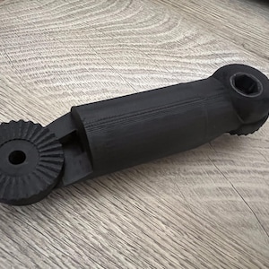 May include: A matte black 3D printed mechanical component featuring a central cylindrical body connected to a circular gear-like end with a central hole and a secondary end with a hexagonal opening, showing visible layer lines throughout.