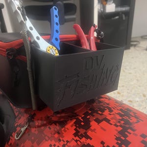 Puede incluir: Un organizador de almacenamiento rectangular de plástico negro con dos compartimentos y el texto DV FISHING en relieve en la parte frontal. Está sujeto a una bolsa negra y roja de la marca HART. El organizador contiene una herramienta blanca y azul y unos alicates rojos.