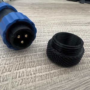 May include: A black and blue three-pin electrical connector with a threaded locking collar and a separate black, textured, cylindrical threaded cap positioned next to it on a light-colored surface.