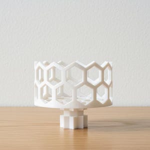 May include: A white, 3D-printed bowl featuring a repeating hexagonal honeycomb pattern throughout its cylindrical walls. The bowl is supported by a central, multi-faceted pedestal base, creating an open, geometric structure.