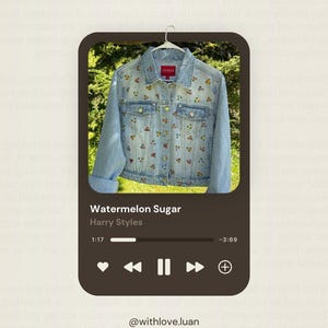 May include: Light blue denim jacket with colorful floral embroidery. The jacket is hanging on a white hanger. The image also includes a music player interface with the song "Watermelon Sugar" by Harry Styles.