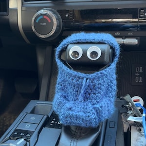 Op de afbeelding: Een blauwe gehaakte hoodie-hoes voor een versnellingspook, voorzien van twee grote wit-zwarte wiebelogen aan de voorzijde. De gebreide stof heeft een textuurpatroon en een klein trekkoorddetail in het midden van de capuchon.