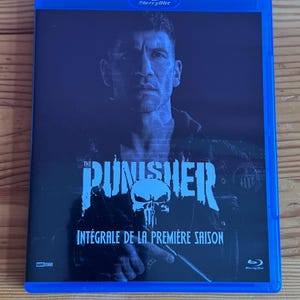 Peut inclure: Un boîtier Blu-ray bleu avec le titre "The Punisher" et un motif de crâne. La couverture présente le visage d'un homme et le texte "Intégrale de la Première Saison". Le boîtier Blu-ray a un bord bleu.