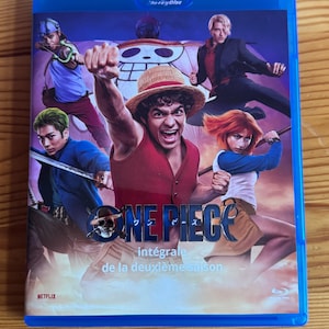 Peut inclure: Un boîtier Blu-ray bleu avec le titre "ONE PIECE" et un logo de tête de mort et d'os croisés. La couverture présente des personnages en action, avec le texte "intégrale de la deuxième saison". Le logo Netflix est visible.