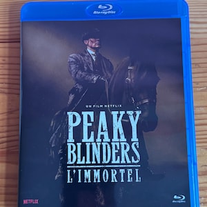 Peut inclure: Un boîtier Blu-ray bleu avec le titre "Peaky Blinders L'Immortel" et un homme à cheval. Le logo Netflix est visible. Le boîtier a une bordure bleue et le logo Blu-ray Disc.