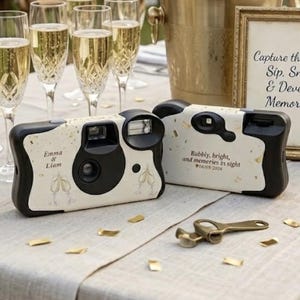 May include: A wedding reception table with champagne glasses, cameras, and a sign saying "Capture the Love: Sip, Snap, & Develop Memories!" Two disposable cameras are decorated with floral designs and the names "Emma & Liam."