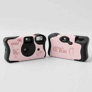 May include: Two pink disposable cameras with black detailing. One camera has the text "BRIDES TO BE" and a bride graphic. The other says "CAPTURE THE BRIDE" with a hand graphic. Wedding theme.