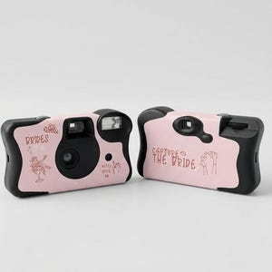 May include: Two pink disposable cameras with black detailing. One camera features the text "BRIDES" and a drawing of a person. The other camera has the text "CAPTURE THE BRIDE" and a drawing of hands. The cameras are for a wedding.