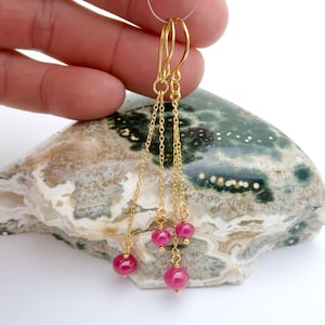 May include: Gold-tone dangle earrings featuring multiple strands of delicate chains. Each chain is adorned with a vibrant, round, pink gemstone. The earrings are suspended from gold-colored ear wires.