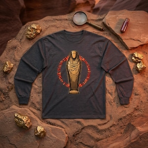 Puede incluir: Una camiseta de manga larga gris carbón con un gráfico central de un sarcófago egipcio dorado adornado con el Ojo de Horus, rodeado por un anillo circular rojo brillante de símbolos jeroglíficos impreso en el pecho.