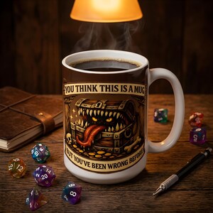 Puede incluir: Una taza de cerámica blanca llena de líquido oscuro, con un gráfico de un cofre del tesoro de madera con dientes afilados y una lengua larga. El texto dice: YOU THINK THIS IS A MUG BUT YOU'VE BEEN WRONG BEFORE. La taza está rodeada de dados poliédricos.