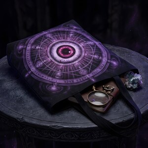 May include: A black tote bag featuring a large, circular purple graphic of a central eye surrounded by intricate geometric patterns, symbols, and the text "THE VOID STARES BACK." A magnifying glass and a small crystal are visible inside the bag.