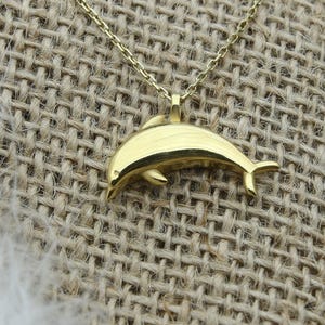 May include: A gold-colored dolphin pendant necklace. The dolphin charm is suspended from a delicate chain. The necklace is displayed on a burlap surface, with a white feather, a pearl bracelet, and a white flower in the background.