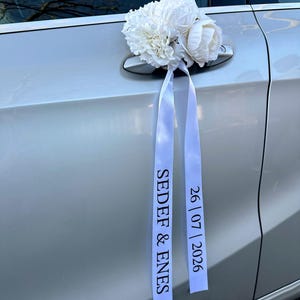 Könnte beinhalten: Ein weißes Blumenarrangement an einem silbernen Autotürgriff mit zwei langen, herabhängenden weißen Bändern. Ein Band trägt den schwarzen Aufdruck SEDEF & ENES, das andere Band zeigt das Datum 26 | 07 | 2026 in schwarzer Schrift.