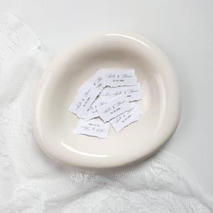 Könnte beinhalten: Eine Sammlung kleiner, rechteckiger weißer Papierstreifen mit gerissenen Kanten, die jeweils mit dem Text "Anita & Haran" und dem Datum "10 | 10 | 2026" in einer schwarzen Schreibschrift bedruckt sind, in einer cremeweißen Keramikschale.