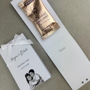 Peut inclure: Un porte-cadeau en carton blanc avec un nœud en ruban blanc, le texte "Heyna & Bilal", "SÖZÜMÜZ SÖZ", "26.07.2026" et une photo en noir et blanc de deux enfants. Un sachet de 6g de café turc Kurukahveci Mehmet Efendi est fixé dessus.