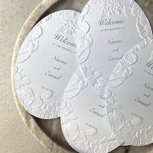 Może przedstawiać: Trzy owalne, białe karty powitalne na ślub z tłoczonymi wzorami kwiatowymi na krawędziach. Tekst brzmi Welcome to the wedding of Naomi and Emsal z datą 30.08.2026 wydrukowaną czarnym tuszem. Karty są ułożone w kształt nakładającego się wachlarza.