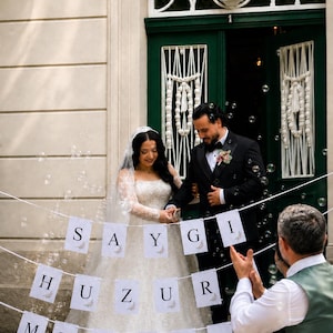 May include: A wedding banner featuring three rows of white rectangular cards with black lettering and floral accents. The top row spells SAYGI, the middle row spells HUZUR, and the bottom row spells MR & MRS, all suspended on white string.