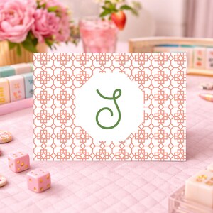 Pode incluir: Um cartão com um padrão geométrico em coral e branco, apresentando uma letra "S" verde dentro de um círculo branco. O cartão está sobre uma superfície rosa, com peças de mahjong e dados no fundo.