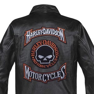 Può includere: Giacca in pelle nera con toppe Harley-Davidson. Le toppe presentano il logo Harley-Davidson, un teschio e le parole "Motorcycles" in lettere arancioni e bianche.