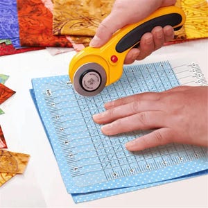 Könnte beinhalten: Ein gelb-schwarzer Rollschneider mit kreisförmiger Klinge auf einem transparenten Patchwork-Lineal mit schwarzen Zoll-Markierungen, platziert auf einem hellblauen Stoff mit weißen Punkten und verschiedenen bunten Stoffresten.