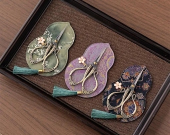 Puede incluir: Tres pares de tijeras de metal ornamentadas con mangos intrincados, cada una con un pequeño dije de flor rosa y una borla de color verde azulado. Las tijeras se exhiben sobre almohadillas de tela con forma de calabaza y patrones variados.