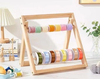 Puede incluir: Un organizador de cintas de madera con dos filas de carretes coloridos. La fila superior presenta cintas pastel, mientras que la fila inferior muestra cintas de satén en varios tonos. El marco de madera natural tiene forma de A.