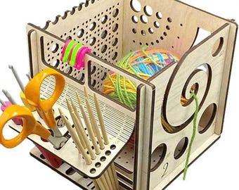 Puede incluir: Un organizador de manualidades de madera con múltiples compartimentos. Contiene hilo, agujas de tejer, tijeras y otros suministros de manualidades. El organizador tiene un acabado de madera clara y presenta cortes decorativos.