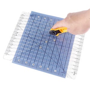 May include: A clear square quilting ruler with black grid lines, inch markings, and diagonal angle guides, positioned over a blue cutting mat. A yellow rotary cutter with a circular blade is placed on the surface of the ruler.