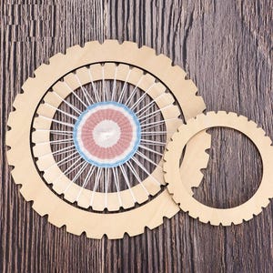 May include: Two wooden weaving looms with a circular design. The larger loom features a woven center with pink, white, and blue yarn. The smaller loom is a separate wooden ring. The background is a dark wood grain.
