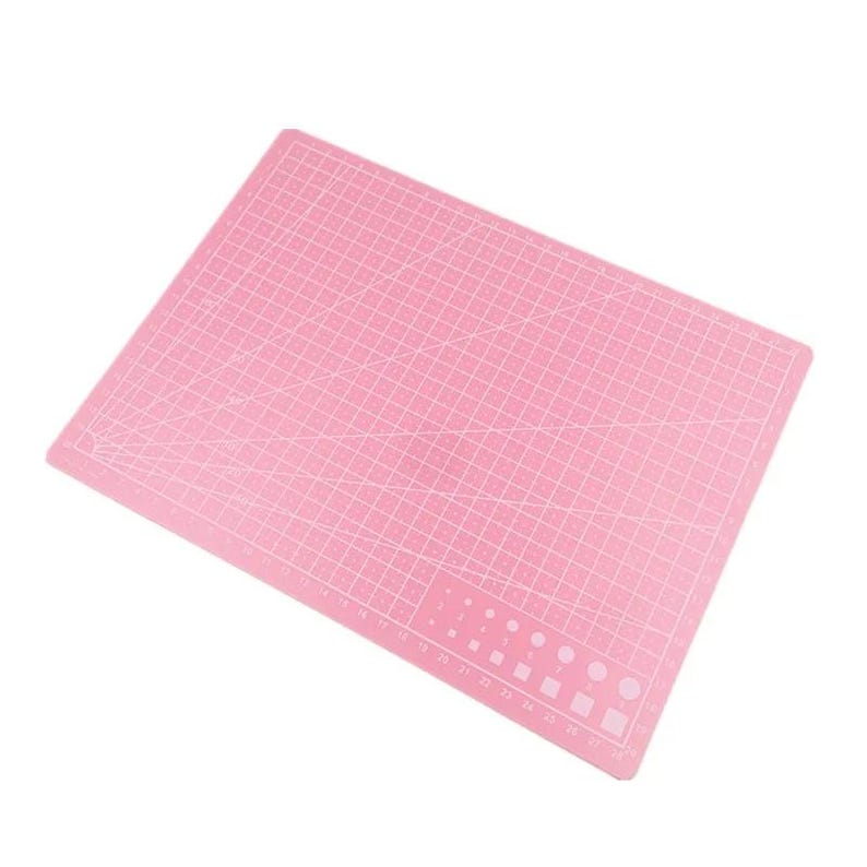 K&ouml;nnte beinhalten: Eine rosa Schneidematte mit einem Rastermuster und Ma&szlig;angaben. Die Matte hat verschiedene Formen und Gr&ouml;&szlig;en f&uuml;r Schneide- und Bastelprojekte. Die Oberfl&auml;che ist f&uuml;r pr&auml;zises Schneiden konzipiert.