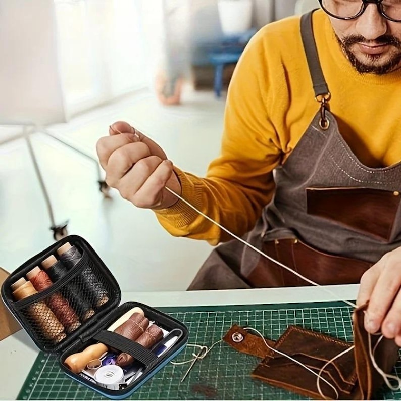 May include: A person working with leather and thread, with a sewing kit and leather pieces on a green cutting mat. The kit contains spools of thread in various colors. The person is wearing a yellow shirt and a brown apron.