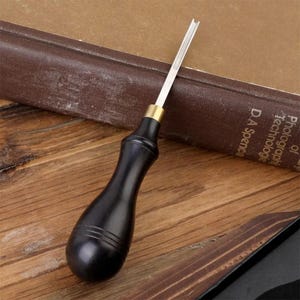 May include: A leathercraft tool with a black wooden handle and a silver blade. The handle has a rounded shape with three grooves near the top. The blade is thin and has a U-shaped tip. A brass collar connects the blade to the handle.