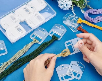 Puede incluir: Un conjunto de bobinas de plástico transparente para hilo de bordar con un estuche rectangular. Varias bobinas están enrolladas con hilos de diversos colores, incluyendo verde claro, verde oscuro, dorado, amarillo y morado.