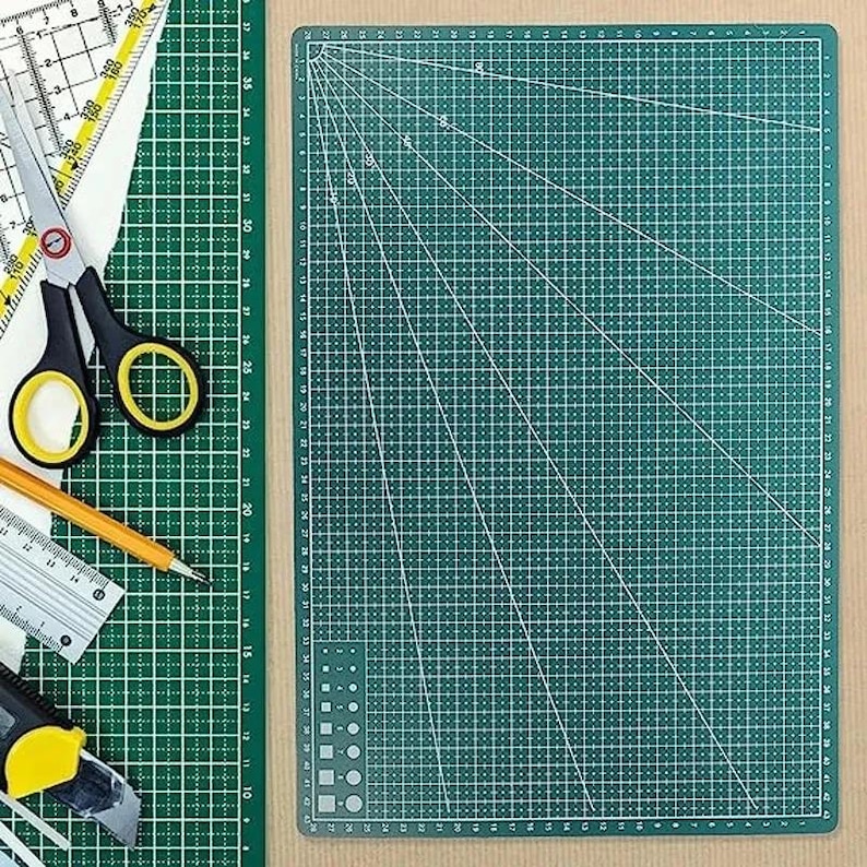 K&ouml;nnte beinhalten: Eine gr&uuml;ne Schneidematte mit wei&szlig;en Gitterlinien und Winkelmarkierungen. Eine schwarze Schere mit gelben Griffen, ein Bleistift, ein Lineal und ein Teppichmesser liegen auf der Matte. Die Matte hat Messungen in Zentimetern.