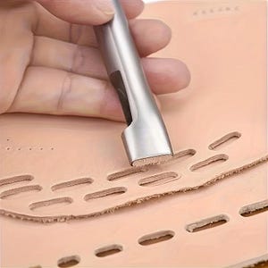 Puede incluir: Se muestra una herramienta de perforación de cuero de metal plateado en uso, creando agujeros oblongos en un trozo de cuero marrón claro. La herramienta es sostenida por una mano. El cuero es de color marrón claro y los agujeros están espaciados uniformemente.