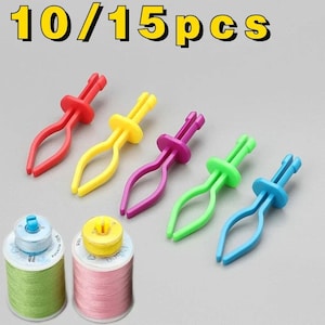 Puede incluir: Un conjunto de cinco clips de costura de plástico en rojo, amarillo, morado, verde y azul, junto a dos carretes de hilo en verde claro y rosa. El texto 10/15pcs aparece en la parte superior de la imagen en letras amarillas con borde negro.