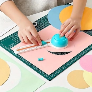 Può includere: Un taglierino circolare bianco e turchese con righello integrato in centimetri, posizionato su un tappetino da taglio verde con griglia. L'utensile presenta un'impugnatura a cupola turchese e un meccanismo a lama per tagliare cerchi di carta.