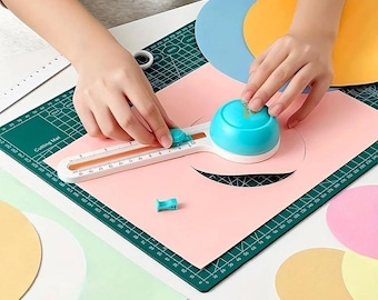 Puede incluir: Un cortador de papel circular blanco y turquesa con una regla integrada marcada en centímetros, sobre una base de corte verde con cuadrícula. La herramienta cuenta con un mango turquesa en forma de cúpula y una cuchilla para cortar círculos.