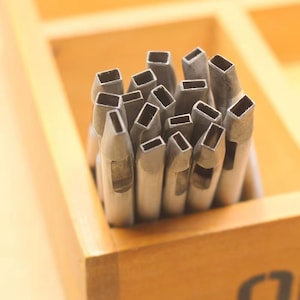 May include: A collection of silver-colored metal rectangular hole punches with hollow tips, arranged vertically in a cluster. Each tool features a distinct rectangular opening at the top, designed for creating precise shapes in various materials.