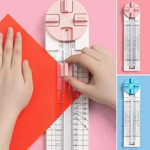 May include: A paper trimmer featuring a white base with a clear ruler and a pink circular dial at the top. The dial has four distinct blade settings for different cuts. The trimmer includes a sliding pink handle for cutting paper.