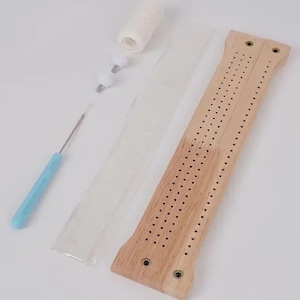 May include: A light-colored wooden cribbage board with three parallel rows of holes, a metal crochet hook with a light blue handle, two small white plastic components, and a white spool of thread, all arranged on a plain white surface.