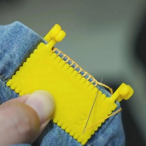 Könnte beinhalten: Ein gelbes Kunststoff-Nähwerkzeug wird zur Reparatur von Denim verwendet. Das Werkzeug ist rechteckig mit einer Reihe von Zähnen an den Kanten. Eine Nadel und Faden sind sichtbar, die den Denim-Stoff nähen. Das Werkzeug wird von einer Hand gehalten.