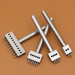 May include: A collection of stainless steel leathercraft tools. The tools have rectangular heads with evenly spaced tines and circular holes. The handles are cylindrical. The tools are arranged on a brown surface.