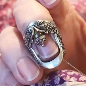 May include: Silver-toned ring featuring an acorn design. The ring has an open design with an acorn in the center, surrounded by detailed leaf and vine patterns. The ring is worn on a finger.