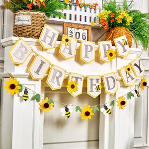 May include: A yellow and white "Happy Birthday" banner with glitter lettering, sunflowers, and bee cutouts. The banner is strung on twine and is decorated with paper sunflowers and bees. The banner is a festive decoration for a birthday party.