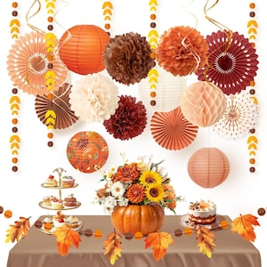 Puede incluir: Un conjunto de decoraciones de fiesta con temática otoñal que incluye farolillos de papel, bolas de nido de abeja y abanicos plisados en naranja, marrón y crema. Un centro de mesa de calabaza con girasoles y un expositor de pasteles.