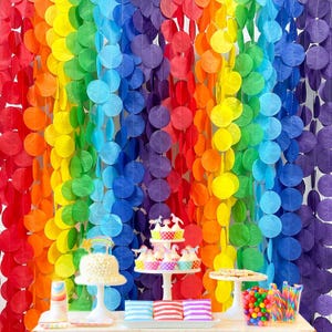 Könnte beinhalten: Eine Party-Szene im Regenbogen-Stil. Ein Hintergrund aus hängenden Papierkreisen in Rot, Orange, Gelb, Grün, Blau und Lila. Kuchen, Cupcakes und Süßigkeiten sind auf einem weißen Tisch angeordnet.